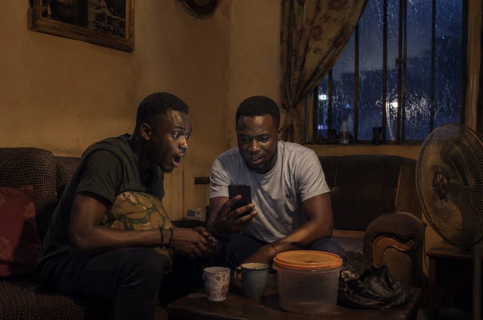The image shows two men sitting together at a table with a cell phone between them. One man appears surprised, while the other looks focused on his phone. The room has a couch against one wall and a fan near another wall. A cup is placed on the table in front of each person. The scene suggests that these men are engaged in conversation or sharing information displayed on their phones.