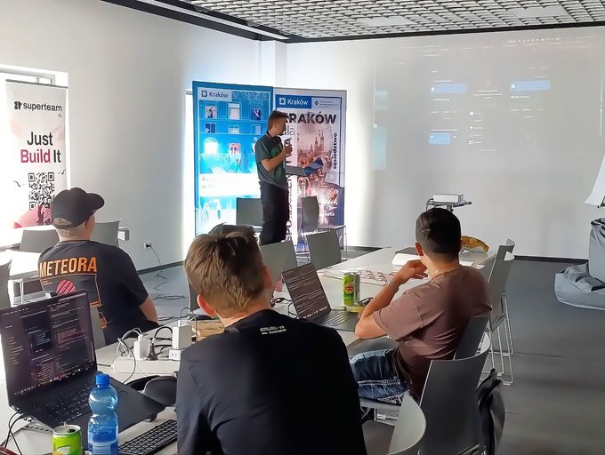 A tech meetup or workshop scene with people working on laptops, "superteam" and "Just Build It" banner, "Kraków" signage, and a presenter at the front.