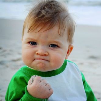 A close-up photo of a young baby with light brown hair wearing a green and white shirt, holding a clenched fist up while standing on a sandy beach with a skeptical expression.