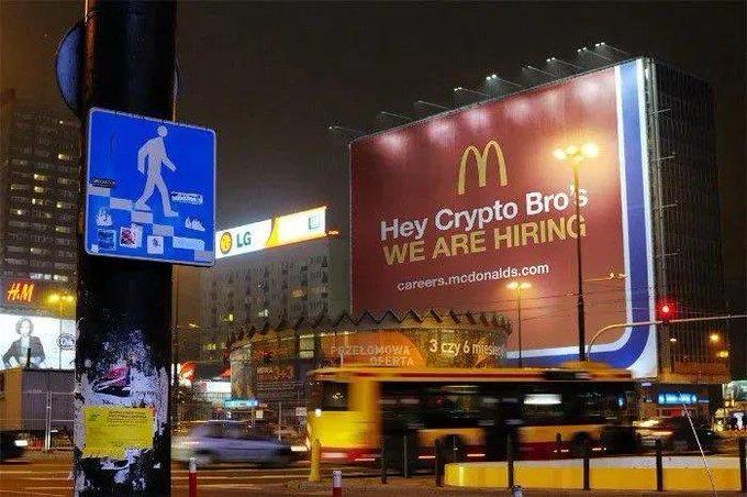 A nighttime street scene featuring a large illuminated McDonald's billboard with the text 'Hey Crypto Bro's WE ARE HIRING' and 'careers.mcdonalds.com' above a passing yellow tram.