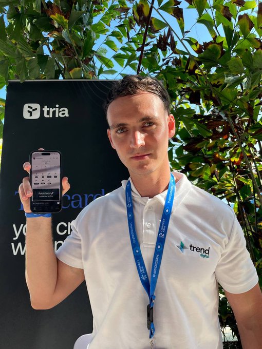 A man wearing a white "trend digital" polo shirt and lanyard holds up his phone displaying the Tria app, standing in front of a branded "tria" backdrop at Coinfest Asia in Bali. The image visually reinforces the post's message about Tria partnering with Trend Digital to showcase their crypto trading app at the event.