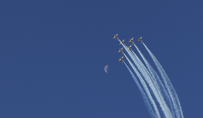 A formation of six jets flies upward in a diagonal line, leaving white contrails against a clear blue sky, with a crescent moon visible nearby.