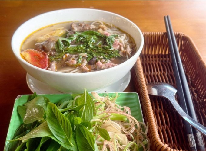 A bowl of Vietnamese noodle soup with beef and greens sits on a wooden table alongside a plate of fresh herbs (basil) and bean sprouts in a woven basket, with chopsticks and a spoon.