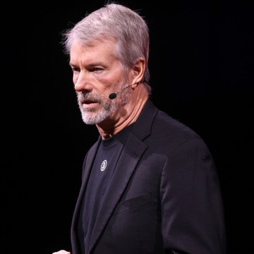 A man with gray hair and a beard, wearing a black blazer and dark shirt, stands on a dark stage wearing a headset microphone while speaking.