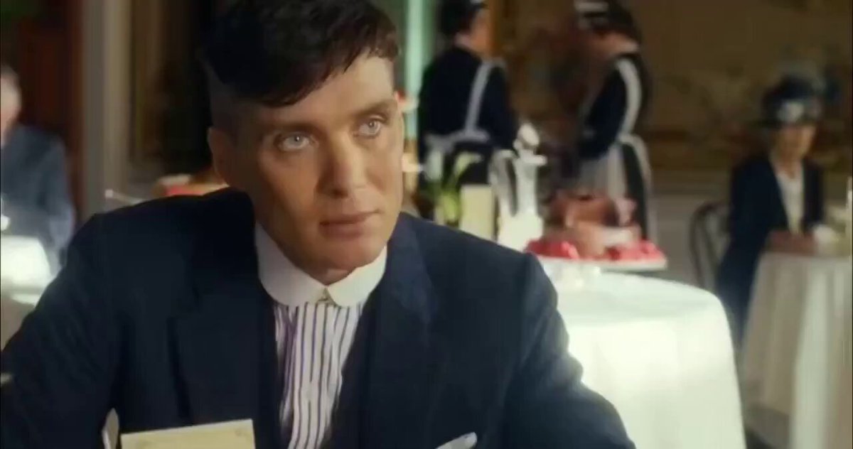 The image shows a man sitting at a table, wearing a suit with a striped tie. The background of the scene includes other people present but not directly involved in the main focus. There are also two cakes visible in the scene, one near the center-left and another towards the right side.