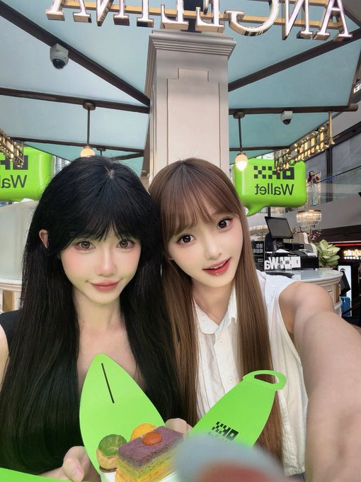 Two young women pose for a selfie inside a Wetzel's Pretzels shop, holding a green paper cone containing a layered cake treat.