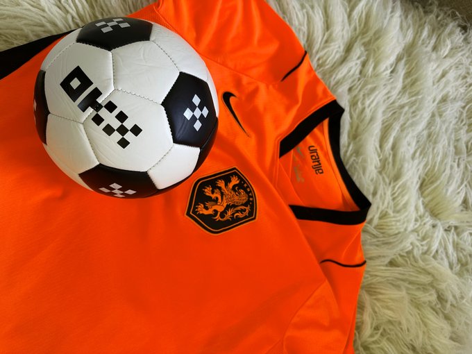The image features a soccer ball, an orange jersey, and a white fur rug laid out on a surface. The soccer ball has black and white checkered designs, while the orange jersey displays a logo of a lion with the word "OKX" written below it. The text in the image reads "@OKX Who are you rooting for?".