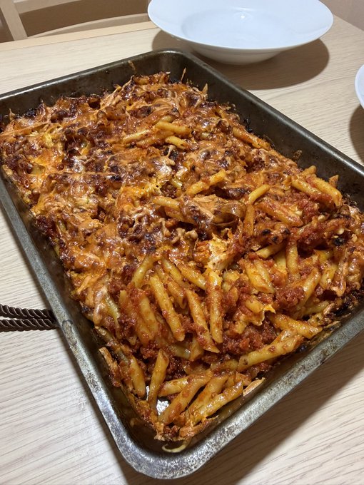 A photograph of a baked pasta dish, appearing to be penne with meat sauce and melted cheese, served in a well-used metal baking pan on a light wooden table, with a white ceramic bowl visible in the background.