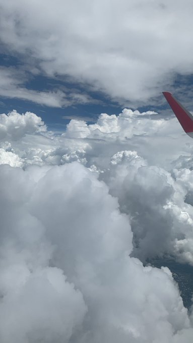 A view from an airplane window showing fluffy white clouds below, with the red-tipped wing of the aircraft visible against a blue sky.