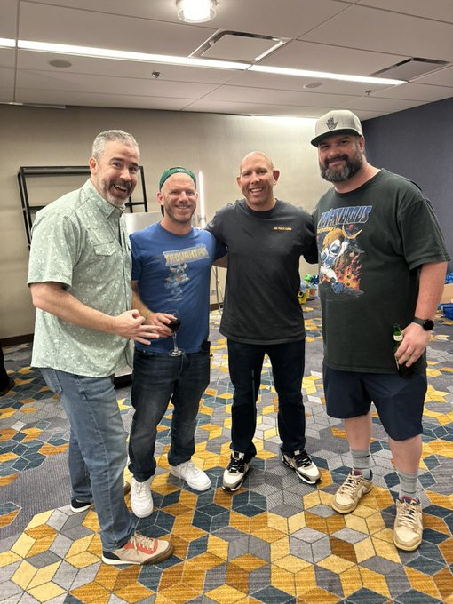 Four men stand smiling together in a room with a patterned carpet; the man on the far right wears a shirt reading 'MASTER BOOT RECORD'.