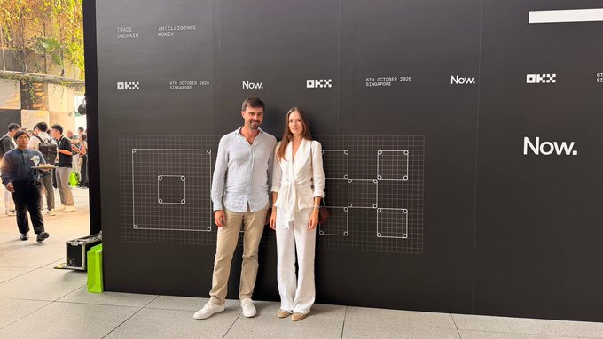 Two people, a man in a light blue shirt and a woman in a white pantsuit, pose together in front of a black backdrop featuring "OKX" and "Now." logos with the date "8TH OCTOBER 2025 SINGAPORE."
