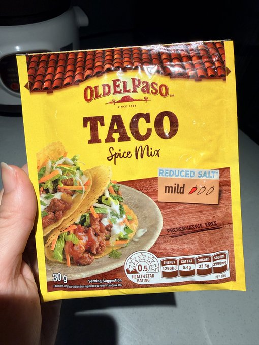 A hand holds up a 30g packet of Old El Paso "Taco" Spice Mix, Reduced Salt and Mild variety, featuring a 0.5 Health Star Rating and 1250kJ per 100g nutritional info on the packaging. The image ties in with the post's humorous Taco Tuesday theme, showcasing the seasoning pack used (or attempted) for taco-making despite the poster's admitted lack of cooking skills.