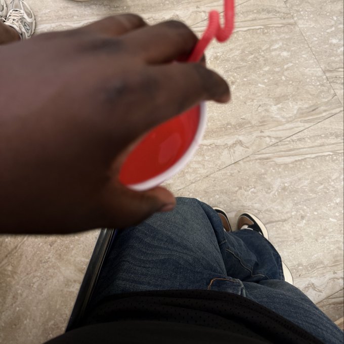 A first-person view looking down at a person wearing jeans and dark sneakers, holding a red plastic cup with a curly red straw over a tiled floor.