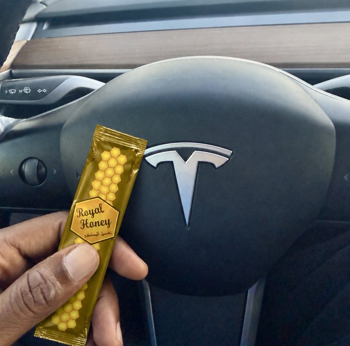 A hand holds a gold 'Royal Honey' packet in front of a Tesla steering wheel with the brand's 'T' logo visible in the background.
