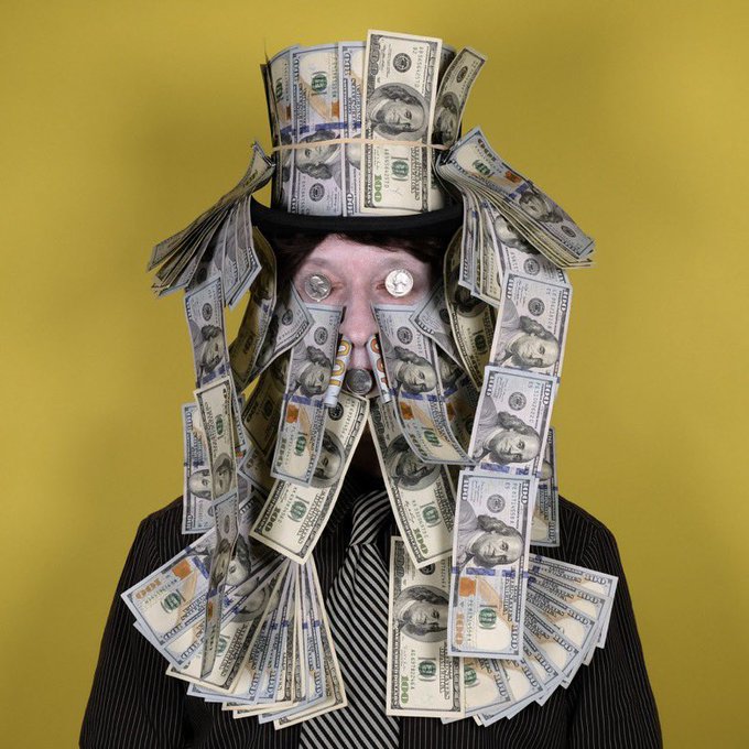 A man wearing a suit, striped tie, and black hat is completely covered in stacks of $100 US bills, with the bills also serving as an elaborate beard and hair decoration against a yellow background. The bills feature Benjamin Franklin's portrait and "100" denominations, making his wealth the central focus of the image.