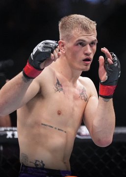 A blonde MMA fighter with chest and torso tattoos stands in a fighting stance wearing black and red MMA gloves inside an octagon cage, likely Ian Garry. The caption context indicates a $20,000 bet was placed on him to win his upcoming Saturday night fight against Islam Makhachev, with a potential payout of $83,560.63 on Polymarket.
