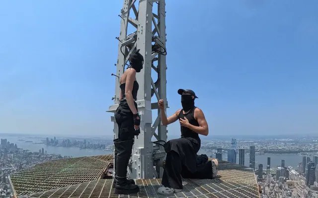 Two people dressed in black with face coverings stand on a high metal tower overlooking a city skyline.