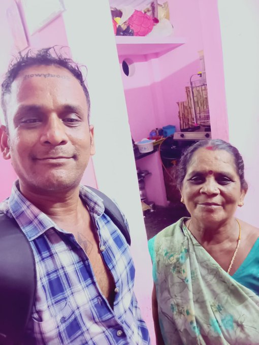 The image features a man wearing a blue and white plaid shirt, standing next to an older woman who is wearing a green sari or shawl. They are both smiling at the camera, creating a warm and friendly atmosphere. The background of the photo shows a pink wall with a shelf on it, which adds to the overall aesthetic of the image.
