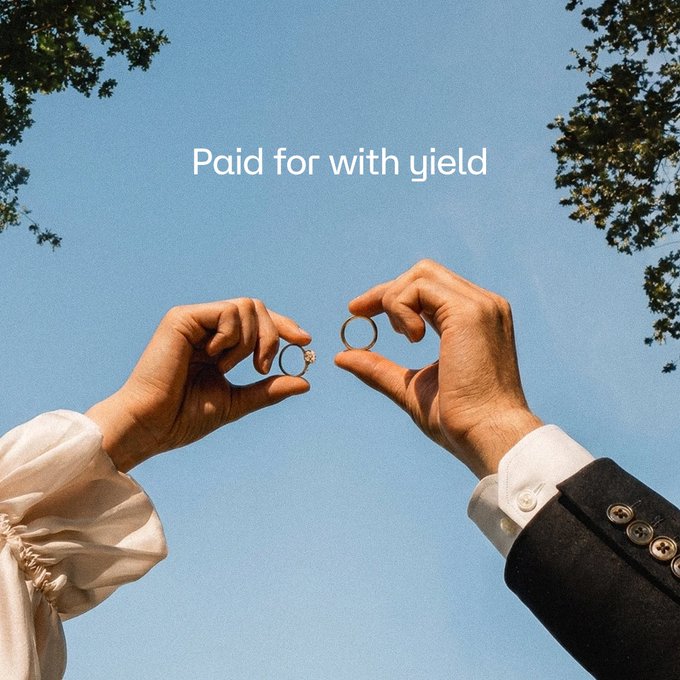 Two hands—one in a delicate cream wedding dress sleeve and the other in a black suit cuff—hold up gold wedding rings against a clear blue sky, while the overlaid text reads "Paid for with yield," illustrating the post's message of using onchain yield from supplying hemiBTC/VUSD via @Vetro_org and @StakeDAOHQ to cover wedding expenses like rings.