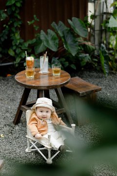A baby in a beige outfit and bucket hat yawns while sitting in a small white camping chair on a gravel patio, with a wooden table behind them holding two glasses of iced coffee and a small pitcher. The scene captures the sleepy "Gm" morning vibe the post describes, playfully contrasting the adult's coffee-fueled readiness to trade with the baby's early-morning fatigue.