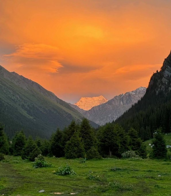 A scenic mountain landscape at sunset, featuring a lush green valley in the foreground and snow-capped peaks bathed in golden-orange light under a hazy sky.