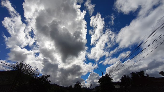 A dramatic sky filled with white and gray clouds against a deep blue background, with power lines stretching across the right side and silhouettes of buildings and vegetation along the bottom.