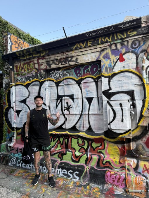 A man in a black tank top, shorts, and a cap stands in front of a heavily graffitied building wall with colorful tags and street art.