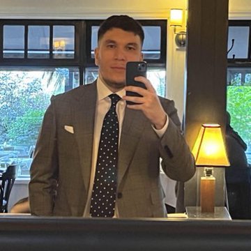 The image features a man dressed in a suit, standing in front of a mirror taking a selfie with his cell phone. The man appears to be wearing a tie with white polka dots. In the background, there are several chairs arranged around dining tables, suggesting that this scene takes place in an indoor setting like a restaurant or a similar venue.