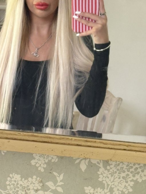 A mirror selfie of a blonde woman wearing a black top, a silver pendant necklace, and holding a phone with a red and white striped case.