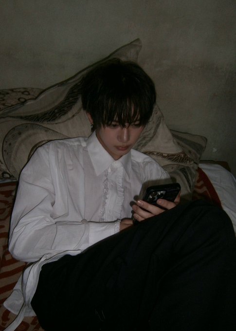 A moody, dimly-lit photo of a young man in a white dress shirt sitting on a bed with patterned pillows, looking down at his phone while covered with a dark blanket.