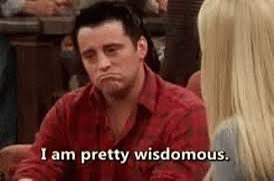 The image features a man wearing a red shirt with the text "I am pretty wise." The man appears to be looking down at his phone, possibly reading or texting. In the background, there are other people present but they are not the main focus of the image.