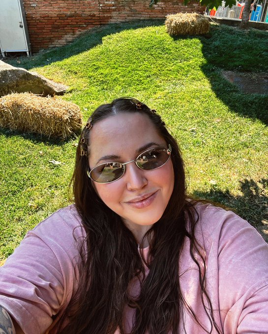 A young woman wearing small oval sunglasses and a pink sweatshirt takes a selfie in a grassy yard with hay bales and a brick building behind her.