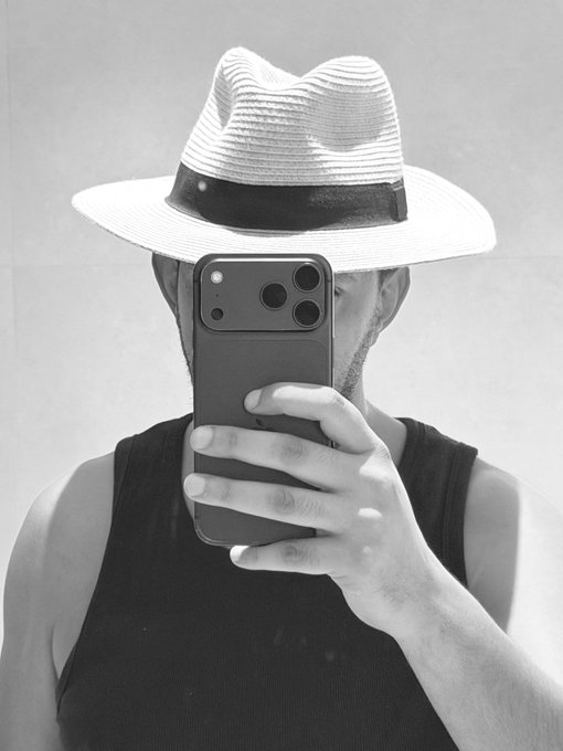 A black and white photo of a person wearing a fedora-style hat and a dark tank top, holding a phone (iPhone 12 Pro) up to their face as if taking a mirror selfie, with the hat's brim fully covering their eyes—mirroring CZ's iconic look in support of the #CZWIFHAT trend.