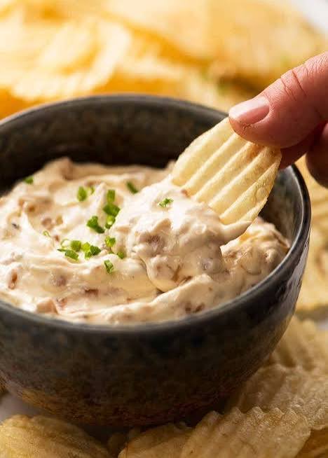 A hand dips a ridged potato chip into a dark ceramic bowl filled with a creamy, onion-flecked dip garnished with chopped chives.