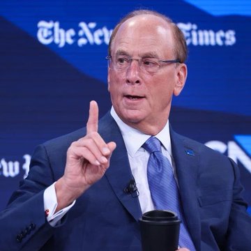 The image features a man dressed in a suit, giving an interview or speaking at a conference. He is holding a cup of coffee while gesturing with his right hand. The background includes a blue wall with white text that reads "The New York Times". The man's position and actions suggest he might be discussing financial matters or sharing insights related to the stock market.