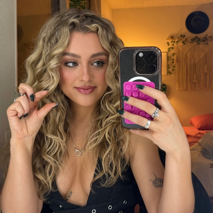 A woman with long curly blonde hair takes a mirror selfie while holding a phone with a pink Pop It case and pointing with her other hand.