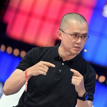 A man with glasses and a shaved head, identified by the post as CZ (Changpeng Zhao), founder of Binance, gestures while speaking on stage during an interview or panel discussion. The accompanying caption references his comments advising traditional finance to adopt crypto to avoid being threatened by it.