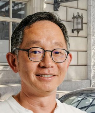 The image features a man wearing glasses and smiling at the camera. He has short hair and is dressed in a white shirt. The background includes a building with large windows and a car parked nearby. There are also two other people partially visible in the scene, but they do not appear to be the main focus of the image.
