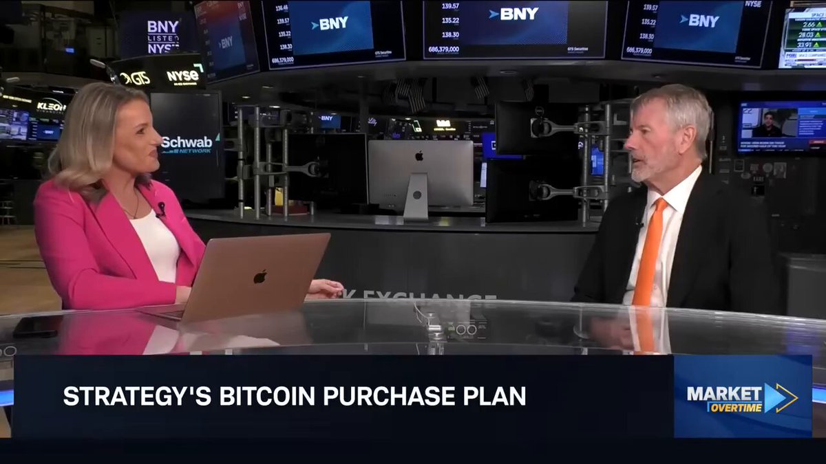 The image shows a man and woman sitting at a table with laptops open in front of them. They appear to be engaged in an interview or discussion about Bitcoin and its purchase plan. The woman has her laptop open, while the man has his laptop closed. Both individuals are wearing ties, indicating a professional setting for their conversation.