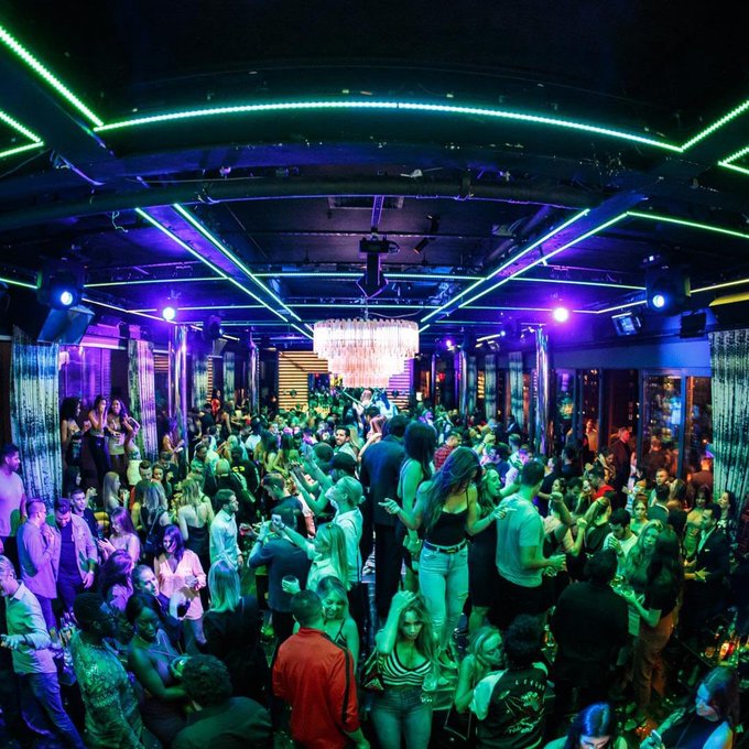 A vibrant nightclub scene with a crowded dance floor illuminated by purple and blue neon lights, featuring a large chandelier hanging from the ceiling as patrons dance and socialize.