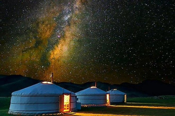A scenic photograph of three illuminated yurts on a grassy plain at night under a brilliant starry sky with the Milky Way visible, set against silhouetted mountains.