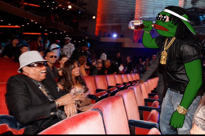 A man in a black suit and hat sits smiling in red theater seats next to a woman, while a green Pepe-style cartoon character wearing a gold chain and cap drinks a beverage in the foreground.