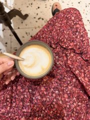 A hand holding a spoon stirring a creamy beverage in a mug, with someone in a red floral dress sitting in the background.