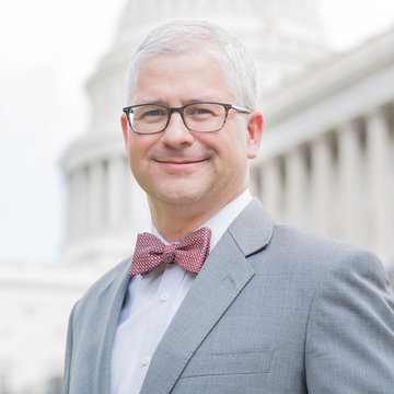 A man wearing a gray suit jacket, light blue shirt, and red-and-white checkered bow tie stands smiling in front of what appears to be the U.S. Capitol building. The image shows no visible text, numbers, or names, and relates to the post simply as a portrait accompanying the word "Correct."