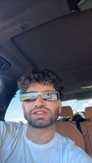 The image features a man wearing sunglasses, sitting inside a car with his head resting against the window. The man appears to be smiling for the camera, possibly posing for a photo or video. The background of the image shows that they are driving on a road, which could suggest an outdoor setting.