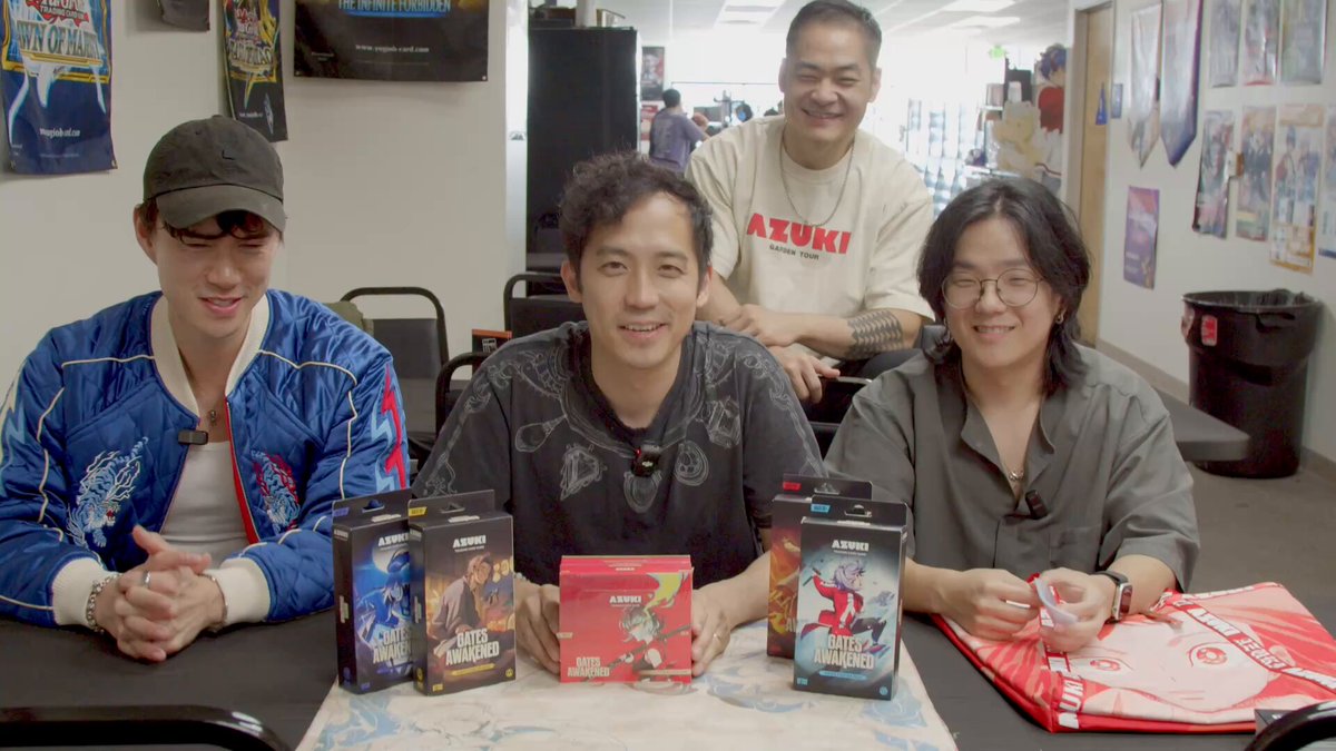 The image shows a group of four people sitting around a table, with three of them holding up boxes containing DVDs or Blu-rays. The fourth person is smiling at the camera while posing for a photo. In the background, there are two chairs visible near the table where they are seated. A TV can be seen in the room as well.