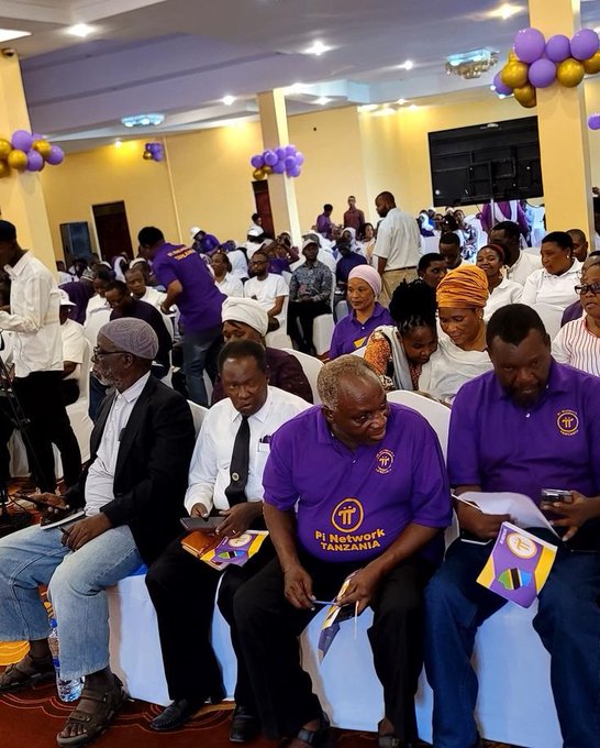 A group of people sit in rows at an indoor gathering, many wearing purple shirts with 'Pi Network TANZANIA' logos, holding Tanzanian flags, with purple and gold balloon decorations visible in the hall.