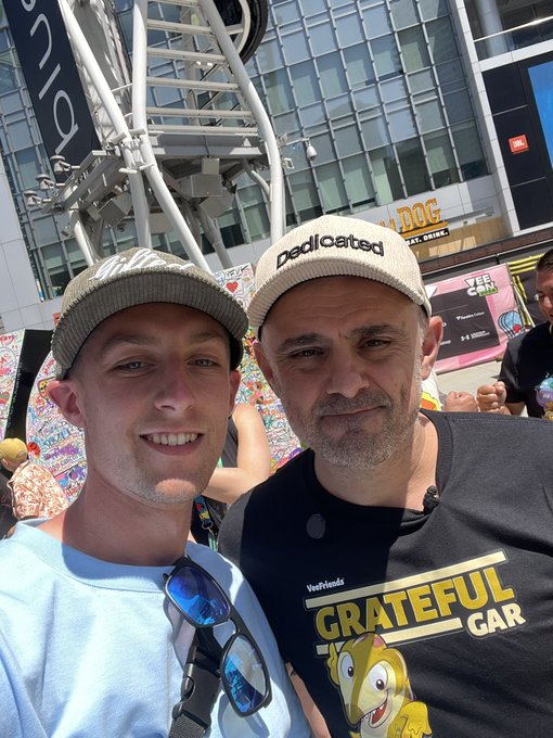 Two men posing for a selfie; one wears a 'pedicated' cap and the other wears a 'Grateful Gar' VeggieFriends shirt, with an event venue in the background.