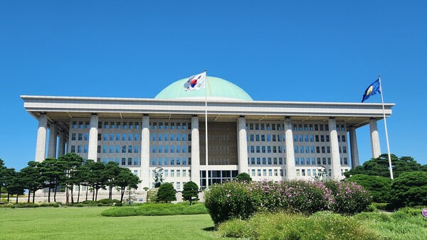 The image shows the South Korean National Assembly building in Seoul, recognizable by its distinctive dome and the South Korean flag flying alongside another banner, serving as the legislative backdrop where People Power Party lawmaker Kim Sang-hoon introduced a bill to delay virtual asset taxation until 2029.