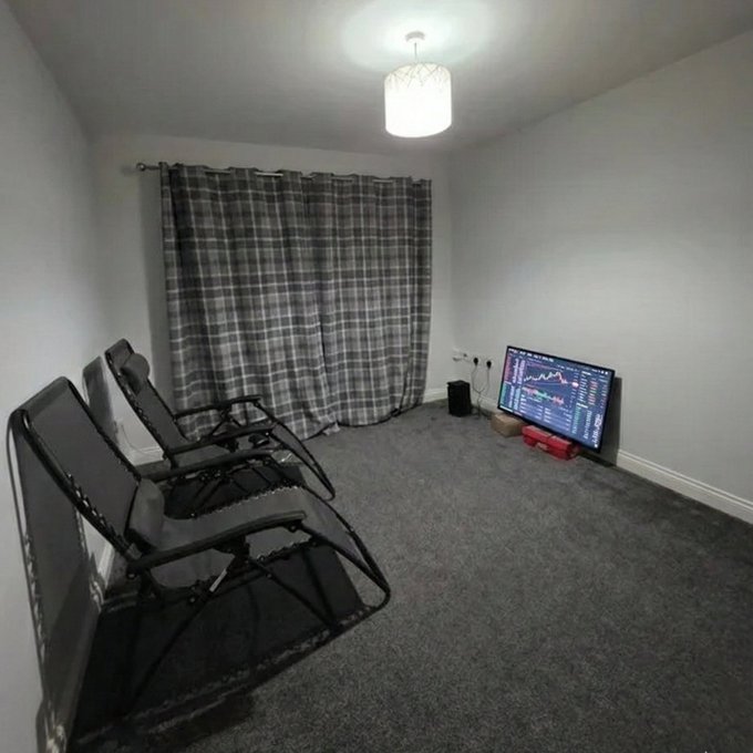 A dimly lit room with gray walls and carpet, featuring a plaid curtain, a ceiling lamp, two black reclining chairs, and a monitor displaying a stock chart on the floor.